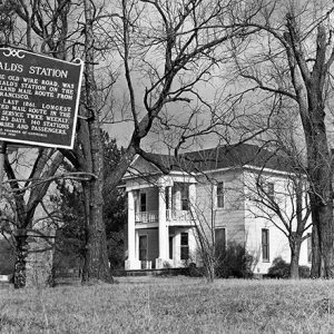 Multistory house with covered porch and balcony under trees with historical marker sign in the foreground