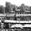 Two white boys fish off wooden bridge with lily pads in foreground, stone lodge in background.