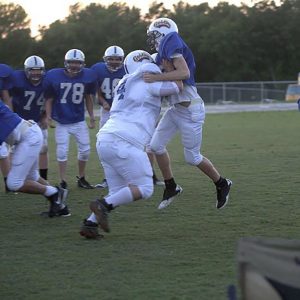A football player pushing a teammate into the air during practice while four individuals play and five teammates observe enthusiastically