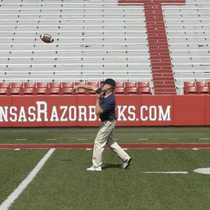 White man on football field in front of stands throwing football into the air