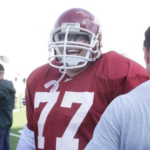 Young white man with glasses football jersey and helmet smiling between two other white men with backs turned